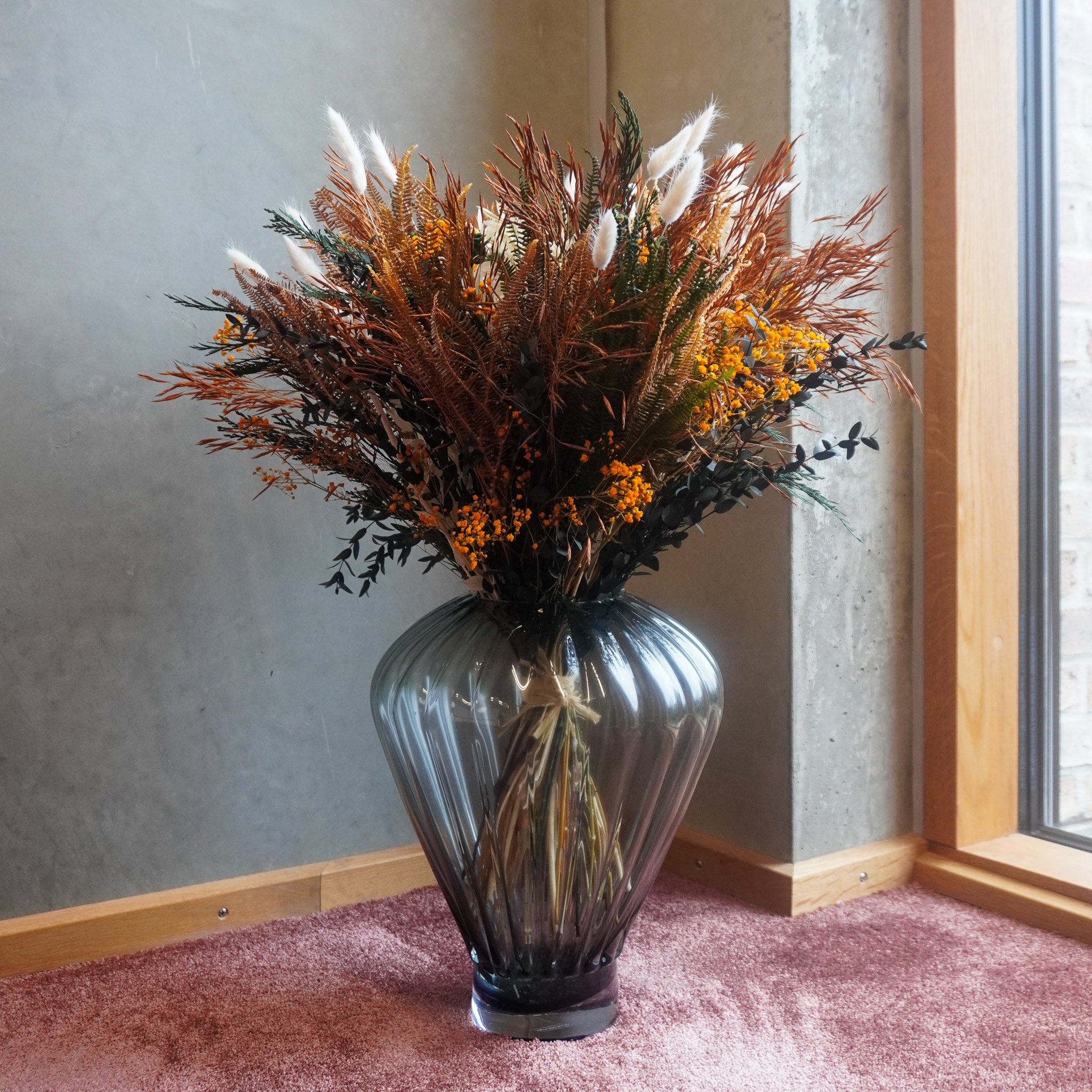I et hjørne af et værelse står en fyldig blomsterbuket i varme efterårsfarver i en geometrisk glasvase på gulvet.