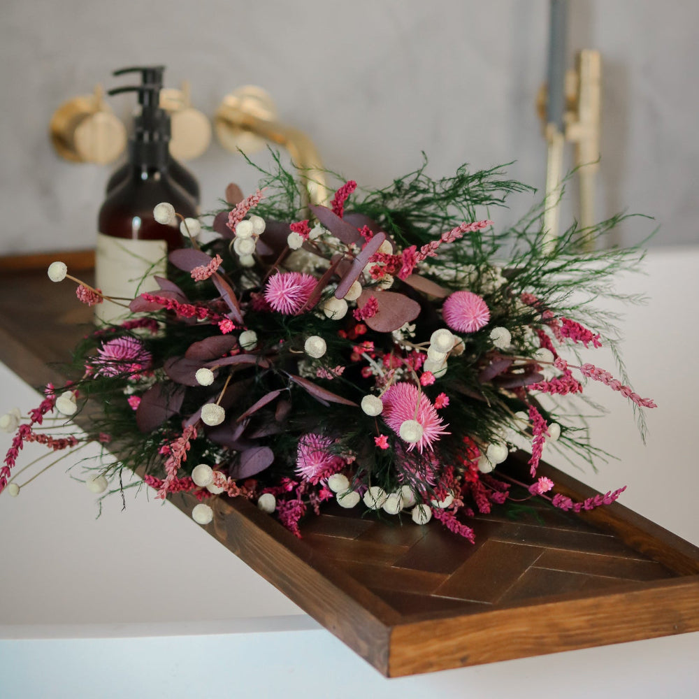 Bukket med langtidsholdbare blomster i pink, lilla og hvide nuancer, placeret på en træbakke ved et badekar.